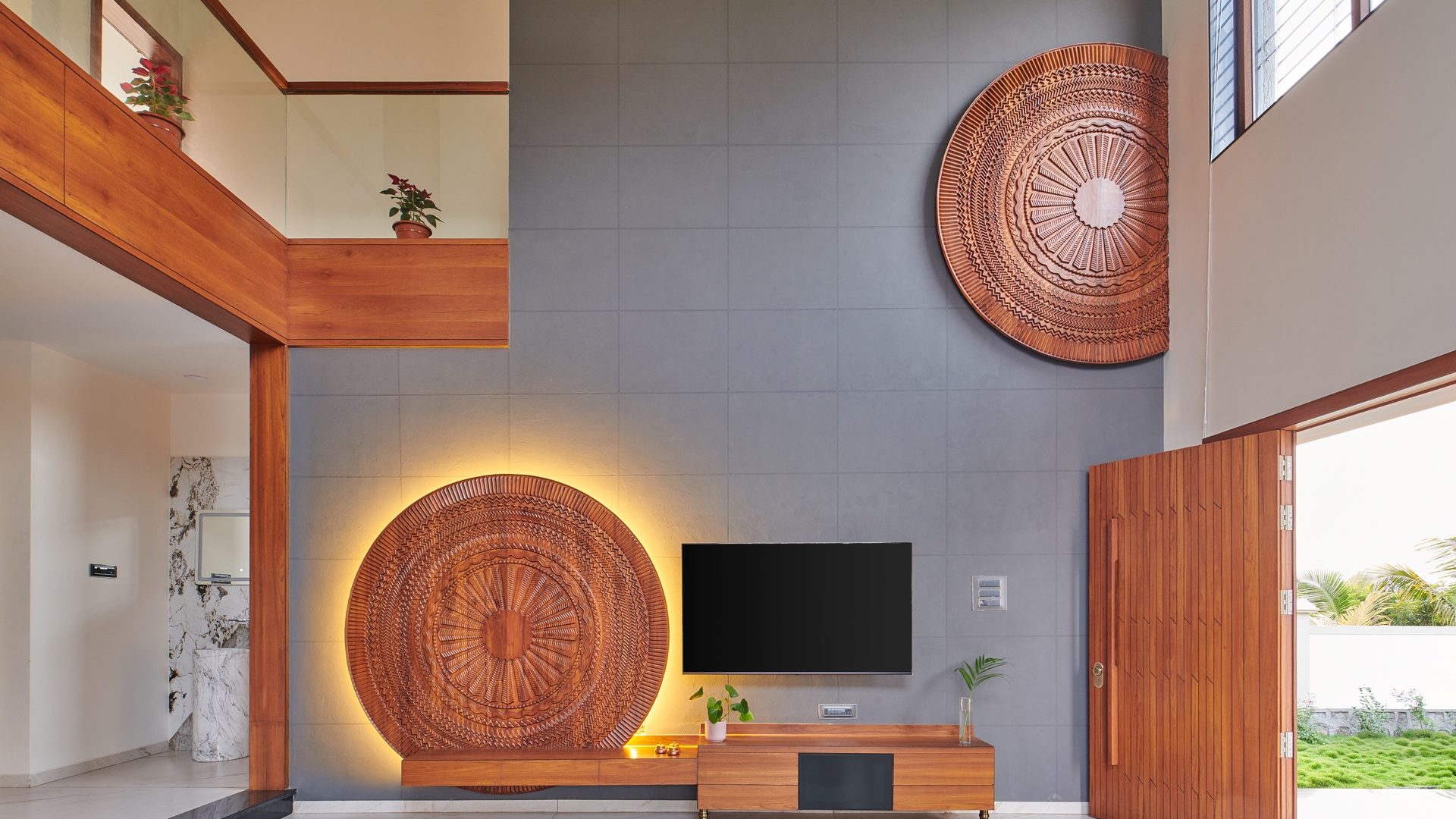 Double-height living room with backlit carved timber medallions on a grey stone wall — Kondhanpuri