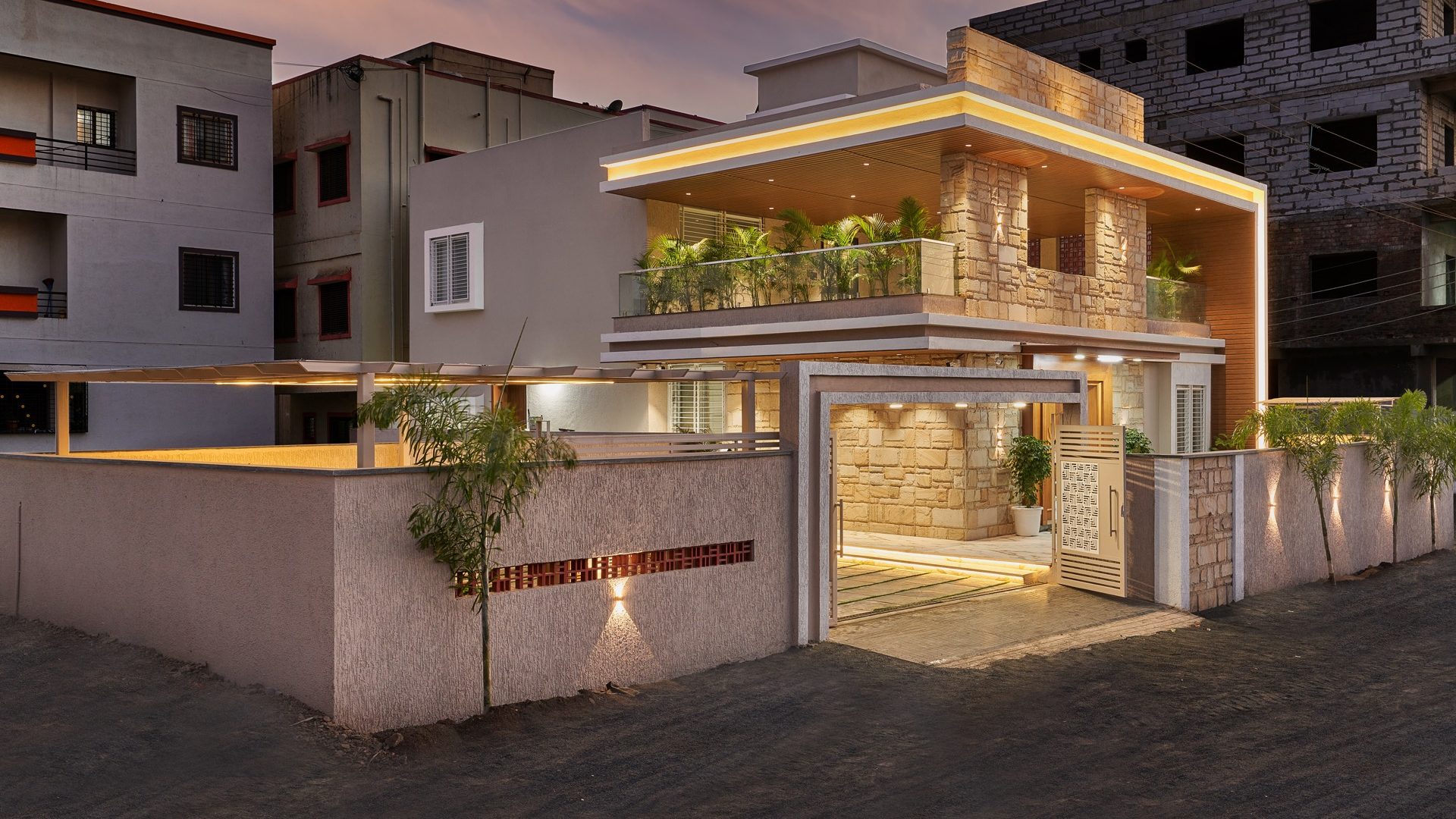 Stone-clad bungalow with a glowing timber soffit and planted balcony at dusk — Nakshatra, Shirur